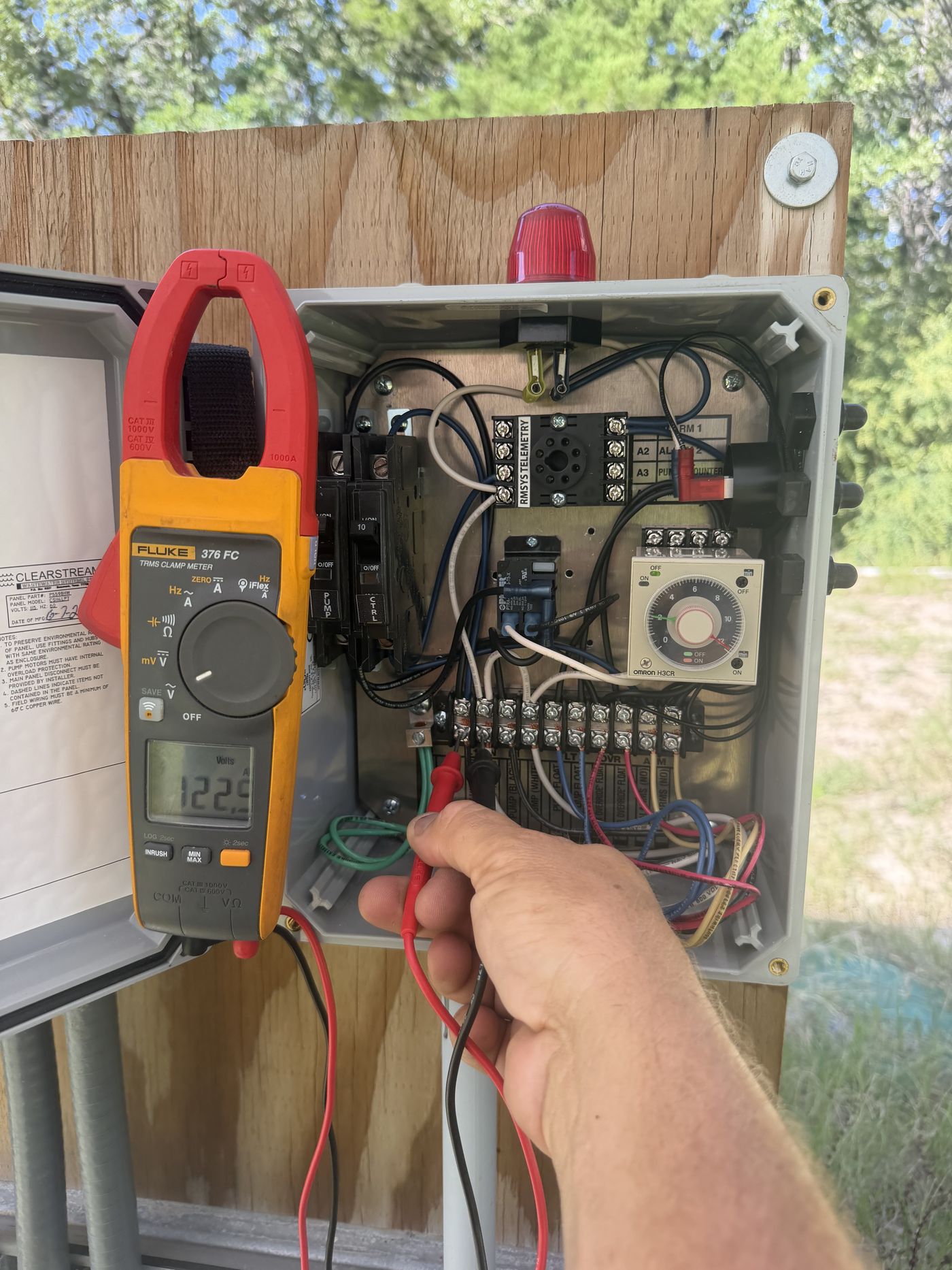 Electrician verifying voltage with a clamp meter inside a pump control panel, reading 122.9 volts